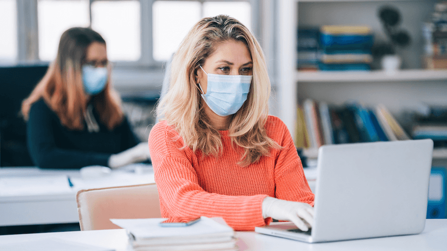 A woman prepares while working on a laptop wearing a surgical mask.